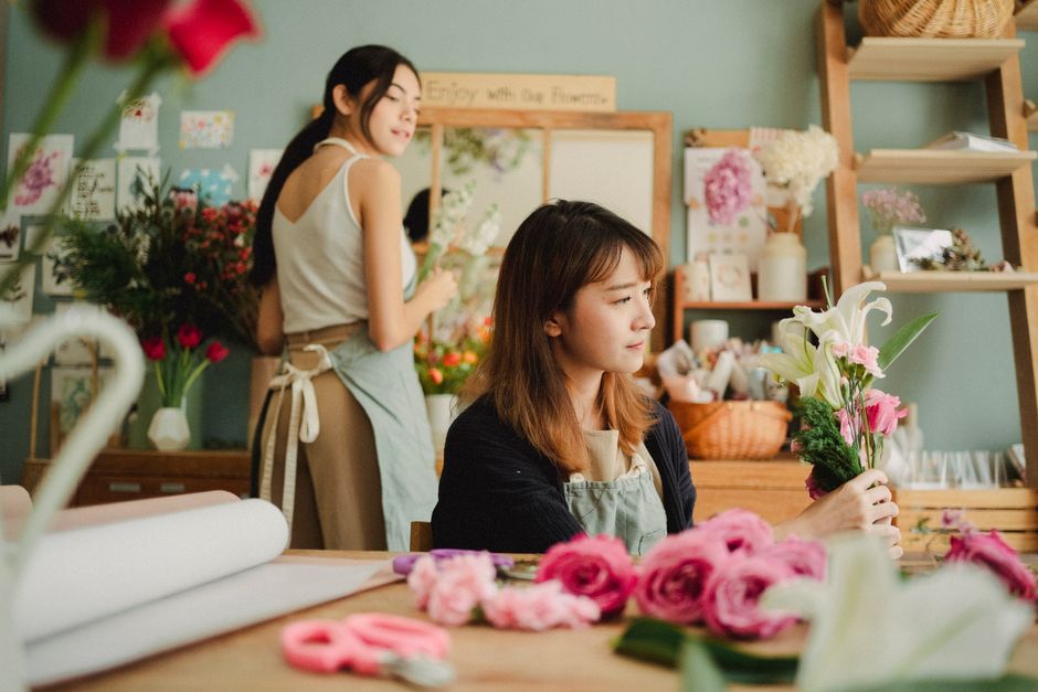 Two women working in a floral workshop, with one woman seated at a wooden table arranging a bouquet of white lilies and pink roses, while the other stands behind her holding a small bouquet. The table